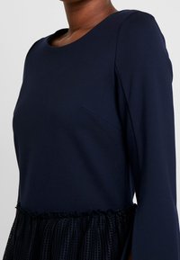 Navy blue dress with a fitted bodice and flared sleeve, featuring a textured ruffled hem and a subtle side seam pocket detail.