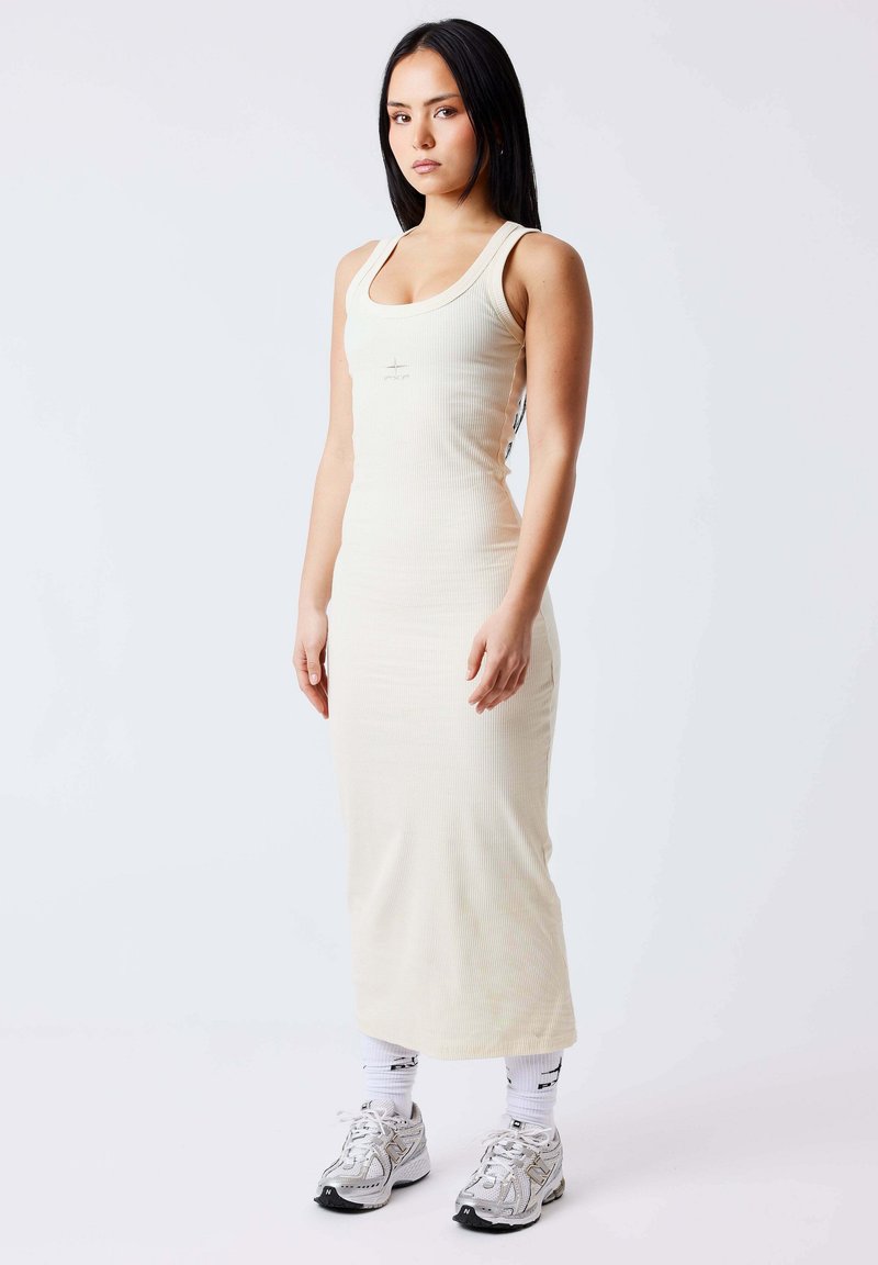 Jeune femme aux cheveux longs noirs portant une robe midi crème sans manches côtelée, des chaussettes blanches et des baskets argentées, debout devant un fond uni.