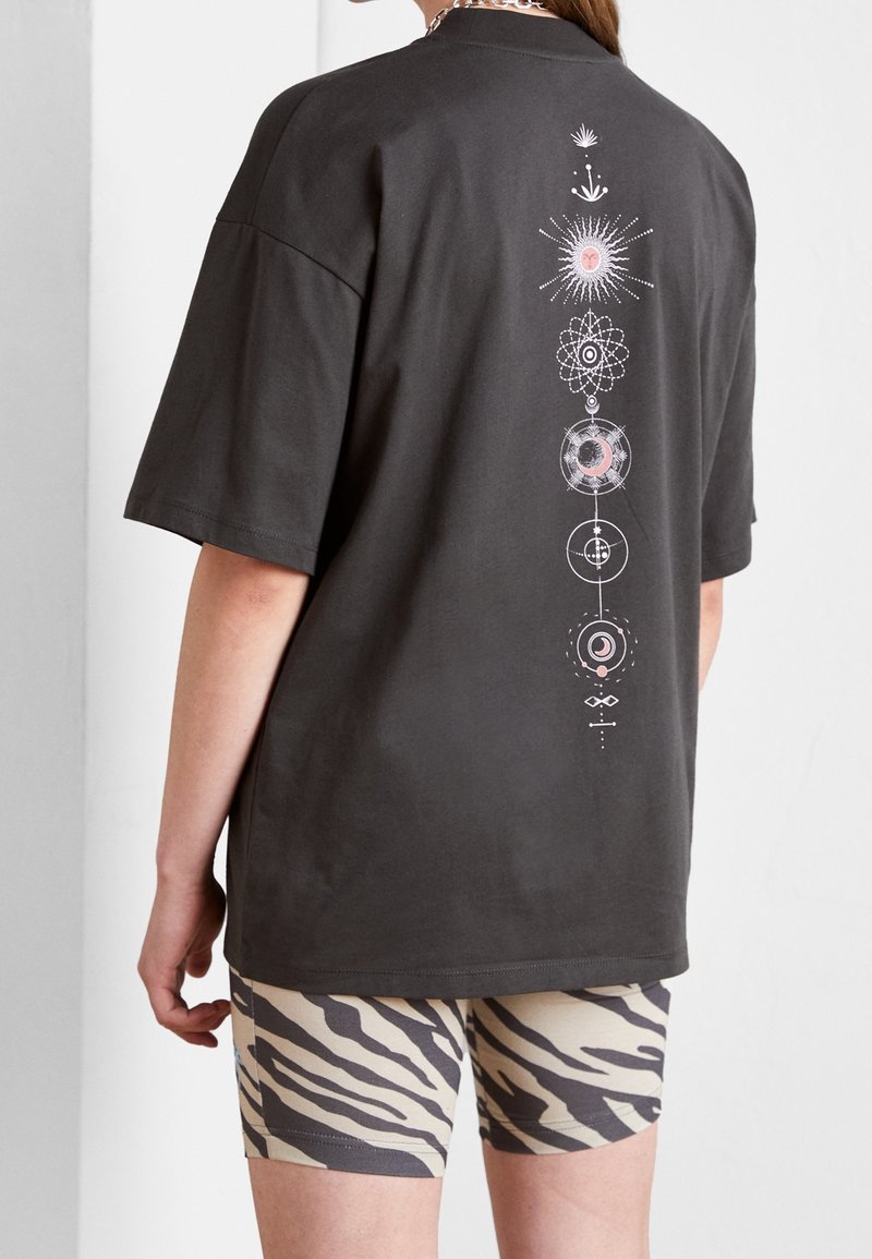 Persona con una camiseta negra de gran tamaño con un diseño celestial vertical en la parte posterior y pantalones cortos de estampado de cebra beige, de pie de lado.