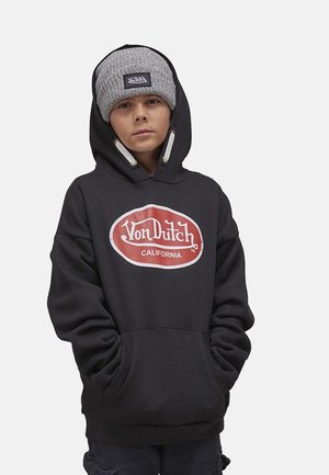 Jeune personne portant un sweat à capuche noir Von Dutch et un bonnet en tricot gris, les mains dans les poches du sweat, sur un fond uni.