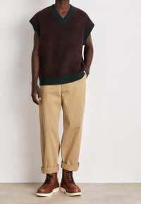 Stickad väst i mörkgrön med röd blommönster, V-hals, korta ärmar; tillsammans med beiga byxor och bruna snörstövlar.