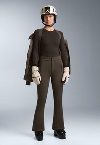 Lyžiarsky outfit pozostávajúci z hnedého priliehavého topu s textúrovaným vzorom, zodpovedajúcich hnedých rozšírených nohavíc, bielych rukavíc a bielej helmy so ski okuliarmi.