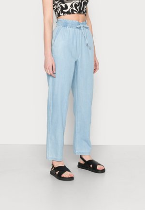 Pantalons bleu clair, amples, avec une taille élastique et un cordon de serrage. Le design décontracté comprend des poches latérales. Portés avec des sandales noires.