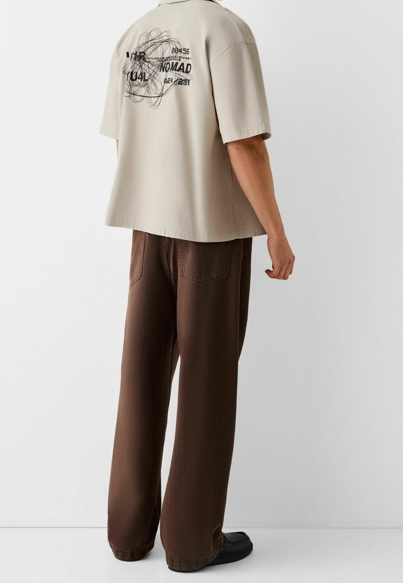 Persona vistiendo una camisa de manga corta beige con texto gráfico negro y pantalones de pierna ancha marrones, de pie frente a un fondo blanco liso.