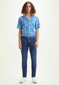 Camisa azul de manga corta con patrones de bandana, combinada con jeans azul oscuro y zapatillas blancas. El conjunto presenta un corte relajado y cierre de botones.