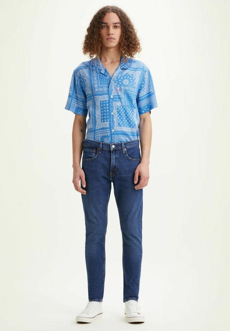 Camisa azul de manga corta con patrones de bandana, combinada con jeans azul oscuro y zapatillas blancas. El conjunto presenta un corte relajado y cierre de botones.