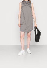 Vestido gris sin mangas con cuello alto, dos bolsillos frontales y un corte relajado. Combinado con zapatillas blancas y sosteniendo un bolso negro.