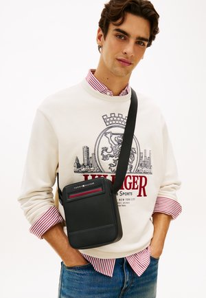 Junger Mann mit weißem Pullover mit roten Streifen, schwarzer Tommy Hilfiger Umhängetasche und blauen Jeans, posiert mit den Händen in den Taschen.