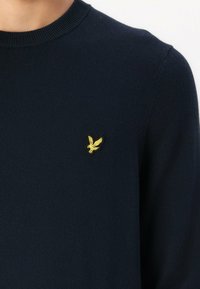 Maglione lavorato a maglia blu navy con scollo rotondo, dotato di un piccolo logo di aquila gialla sulla zona sinistra del petto. Texture liscia, design aderente.