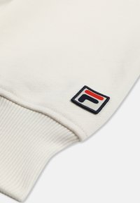 Weißer, gerippter Ärmelbund eines Kleidungsstücks mit einem kleinen rechteckigen Aufnäher, der ein stilisiertes rotes und marineblaues Logo zeigt.