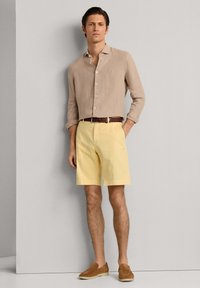 Homme portant une chemise beige à manches longues, un short jaune avec une ceinture marron et des chaussures marron sans lacets, debout contre un mur gris.