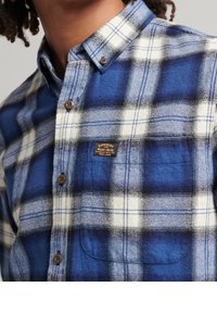 Camicia in flanella a quadri blu e bianca con colletto button-down, taschino sul petto e bottoni marroni; presenta un'etichetta del marchio sulla tasca.