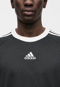 Mörkgrå sporttröja med vit krage och tre vita ränder på axlarna. Har en vit Adidas-logotyp på bröstet.