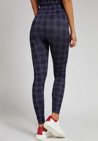 Marineblauwe leggings met een herhalend lichtgrijs logo patroon. Getextureerde stof, aansluitend ontwerp, gecombineerd met rode en witte sportschoenen.