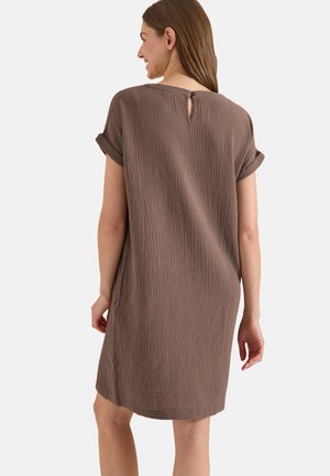 Femme portant une robe marron ample et texturée à manches courtes avec une petite fermeture bouton trou de serrure au dos, debout de dos.