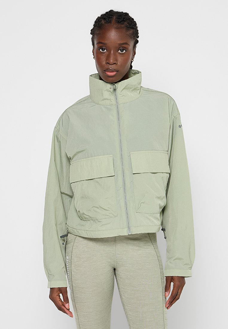 Veste courte vert clair avec un col haut, deux poches avant et une finition lisse et texturée ; fermeture éclair et accents latéraux.