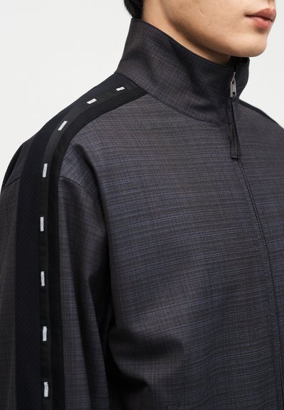 Bărbat purtând o jachetă gri închis cu fermoar, cu guler înalt și detalii negre pe marginea umerilor și a mânecilor.