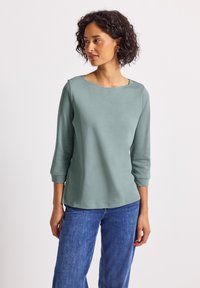 Vrouw met krullend donker haar, gekleed in een lichtgroene lange mouwen top en blauwe spijkerbroek, kijkt naar links tegen een vlakke achtergrond.