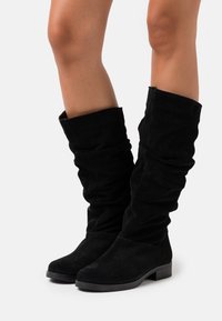 Bottes noires en daim montant jusqu'au genou avec un design flou et un bout arrondi. Talon plat, texture lisse et détails de couture le long des coutures.