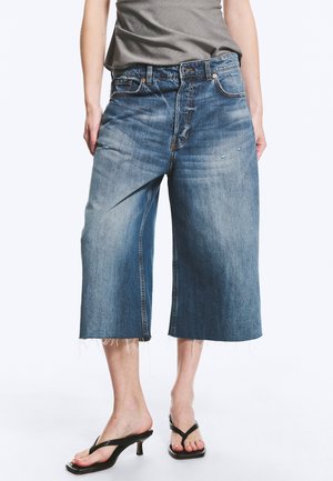 Person trägt weit geschnittene, gekürzte blaue Jeans mit rohen Säumen und schwarze Sandalen mit Absatz und steht vor weißem Hintergrund.