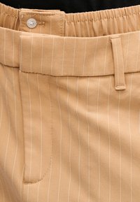 Pantalon beige en tissu texturé, avec fines rayures blanches verticales, taille élastique et une fermeture par un bouton.