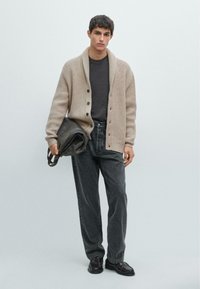 Beiger Rippstrick-Cardigan mit Schalkragen, grauem T-Shirt, dunkelgrauen Jeans und schwarzen Schuhen. Hält eine dunkle Ledertasche mit Tragriemen.