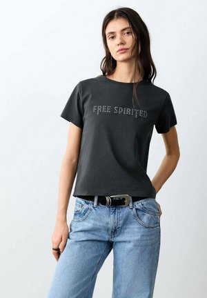 Tânără cu păr lung și închis la culoare, purtând un tricou negru cu ținte și inscripția „Free Spirited” și o pereche de blugi deschiși la culoare, cu o curea neagră.
