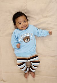 Ljusblå sweatshirt med "LUCKY" och ett björnmotiv; kombinerad med randiga byxor i brunt, blått och krämvitt. Lutar sig mot en beige yta.