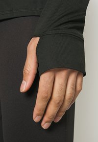 Handen vilar vid sidan iförd en svart långärmad tröja med tumhål och svarta texturerade leggings.