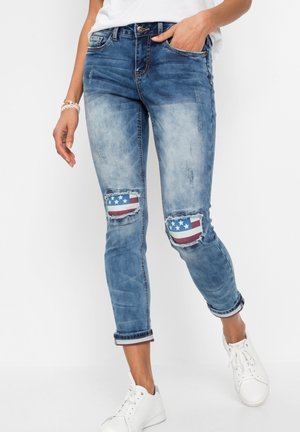 Donna con jeans azzurri sbiaditi con toppa della bandiera americana sulle ginocchia strappate e scarpe da ginnastica bianche, in piedi con una mano in tasca.