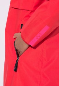 Jaskrawoczerwona wodoodporna kurtka z kieszenią na zamek, ściągaczami na rękawach oraz kontrastowym różowym akcentem z logo na rękawie.