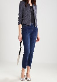 Marineblauwe getextureerde cropped jas, zwart henleytopje, spijkerbroek met rafelige zoom en glanzende zilveren hakken. Houdt een witte en zwarte paraplu vast.