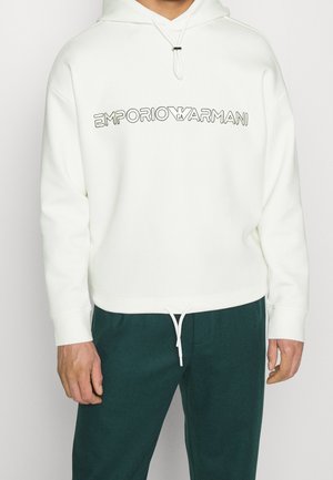 Sudadera blanca con capucha que presenta un estampado del logo en la parte frontal, cierre con cordón y puños acanalados. Combinada con pantalones jogger texturizados en verde oscuro.