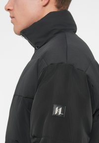 Juodas pufinis švarkas su aukštu apykakle, tekstūruotais peties ir rankovėmis akcentais, kairėje rankovėje esantis prekės ženklo patch.