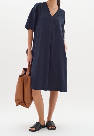Vrouw draagt een losse marineblauwe V-halsjurk, zwarte slidesandalen en houdt een grote bruine totebag vast tegen een effen witte achtergrond.