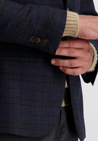 Mann in einem dunkel karierter Blazer über einem beige gestrickten Pullover, mit gefalteten Händen vor ihm, der die Knöpfe und das Detail der Blazerärmel zeigt.