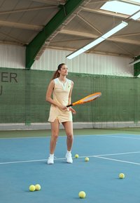 Joueuse de tennis portant une robe sans manches jaune clair avec un col, tenant une raquette orange, sur un court bleu avec plusieurs balles de tennis autour.