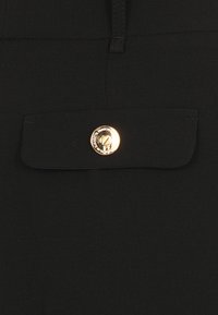 Un tessuto nero presenta una tasca a patta chiusa con un bottone gold circolare. Superficie liscia con leggero splendore, senza motivi o rifiniture visibili.