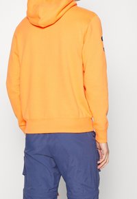 Orange cotton hoodie with a kangaroo pocket, ribbed hem, and cuffs. Worn over navy shorts; hood is pulled up for coverage.