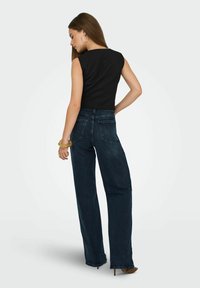 Frau mit langen Haaren, die ein ärmelloses schwarzes Oberteil und dunkle weit geschnittene Jeans trägt, steht mit einer Hand auf der Hüfte, abgewandt.