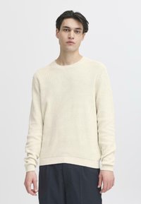 Maglione a maglia color crema con scollatura rotonda, maniche lunghe e texture a coste. Indossato con pantaloni scuri, sfondo minimale.