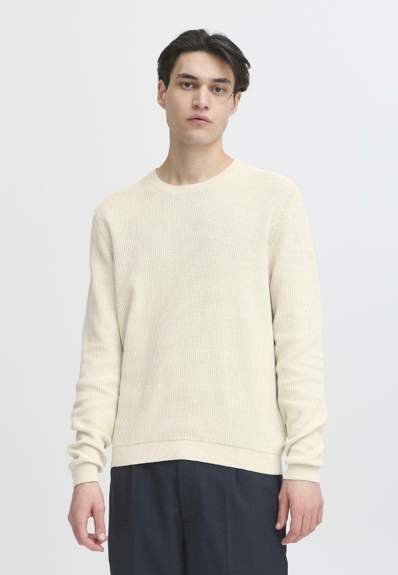 Maglione a maglia color crema con scollatura rotonda, maniche lunghe e texture a coste. Indossato con pantaloni scuri, sfondo minimale.