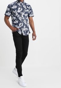 Kortärmad skjorta med knappar, marinblå bakgrund och vitt blommönster, kombinerad med svarta smala jeans och vita sneakers.