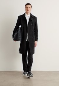 Abrigo trench de doble botonadura negro con un corte ajustado, llevado sobre una camisa blanca, pantalones negros y zapatillas negras, mientras sostiene un bolso negro.