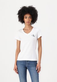 White v-neck t-shirt in soft cotton fabric, featuring short sleeves and a small black logo at the left chest. Paired with blue jeans.
