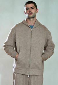 Brun hættetrøje med lynlås, stor hætte, tekstureret stof, forlommer og ribbede manchetter, båret over en blå skjorte, med en afslappet pasform.