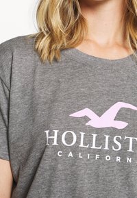 Grå T-shirt i mjukt material med en rosa måslogga och vit text "HOLLISTER CALIFORNIA" på framsidan.