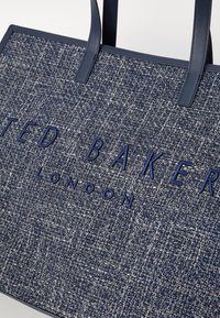 Blauwe geweven tas met leren accenten, met de gegraveerde merknaam "TED BAKER LONDON" aan de voorkant. Rechthoekige vorm, stevige handvaten.