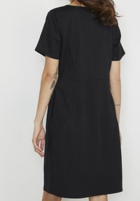 Vestido negro de manga corta, confeccionado en un tejido suave, con un corte holgado, escote modesto, bolsillos laterales y detalles en la costura trasera.