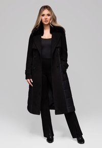 Kvinde med langt blond hår iført en sort pelskravefrakke, sort top, sorte bukser med frontslids og sorte ankelstøvler, stående lige.